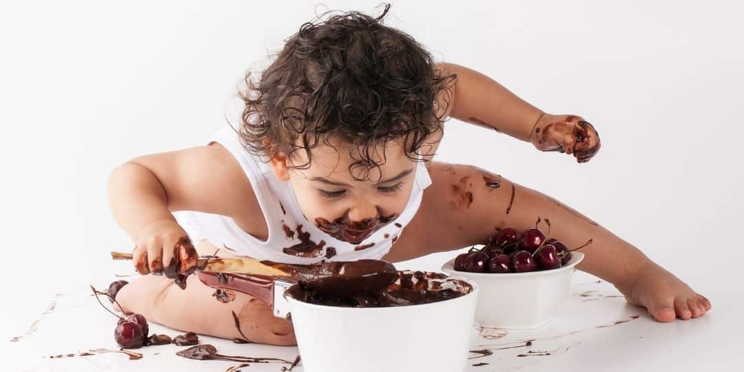 Child covered in chocolate looking into a pot of melted chocolate