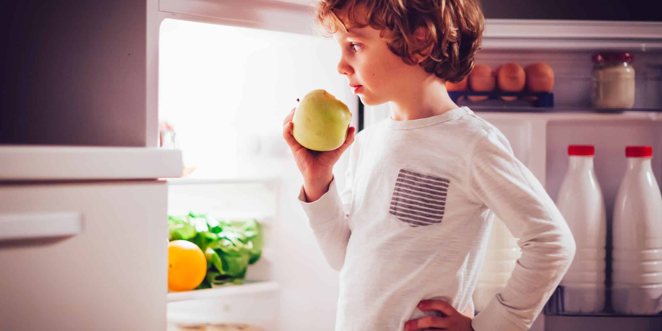 boy having a bedtime snack of an apple in his pajamas standing in the open door of the fridge